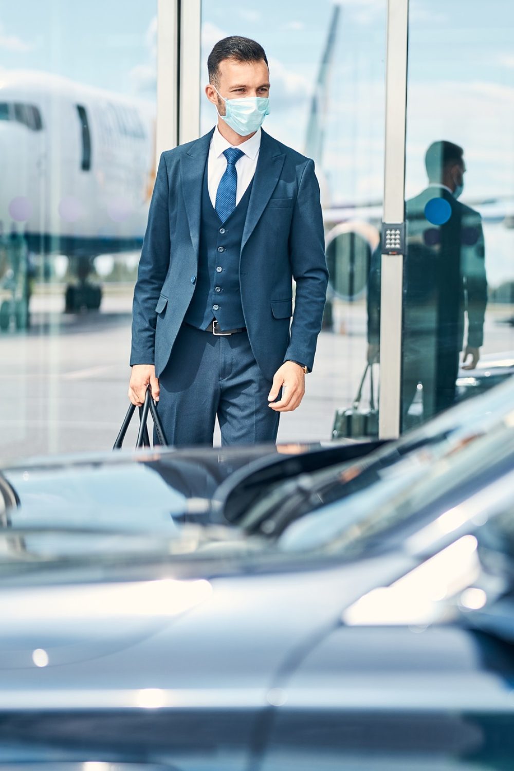 Businessman with a bag in hand standing near the glass door of an airport and looking at the car in front of him. Medical mask on his face